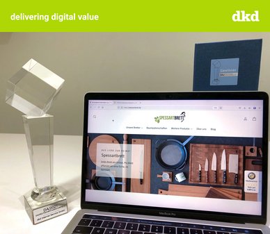 Grüner Balken oberhalb des Bildes mit der Aufschrift delivering digital value und dkd. Es ist der Deutsche Agenturpreispokal zu sehen inklusive MacBook mit der Website von Spessartbrett.com
